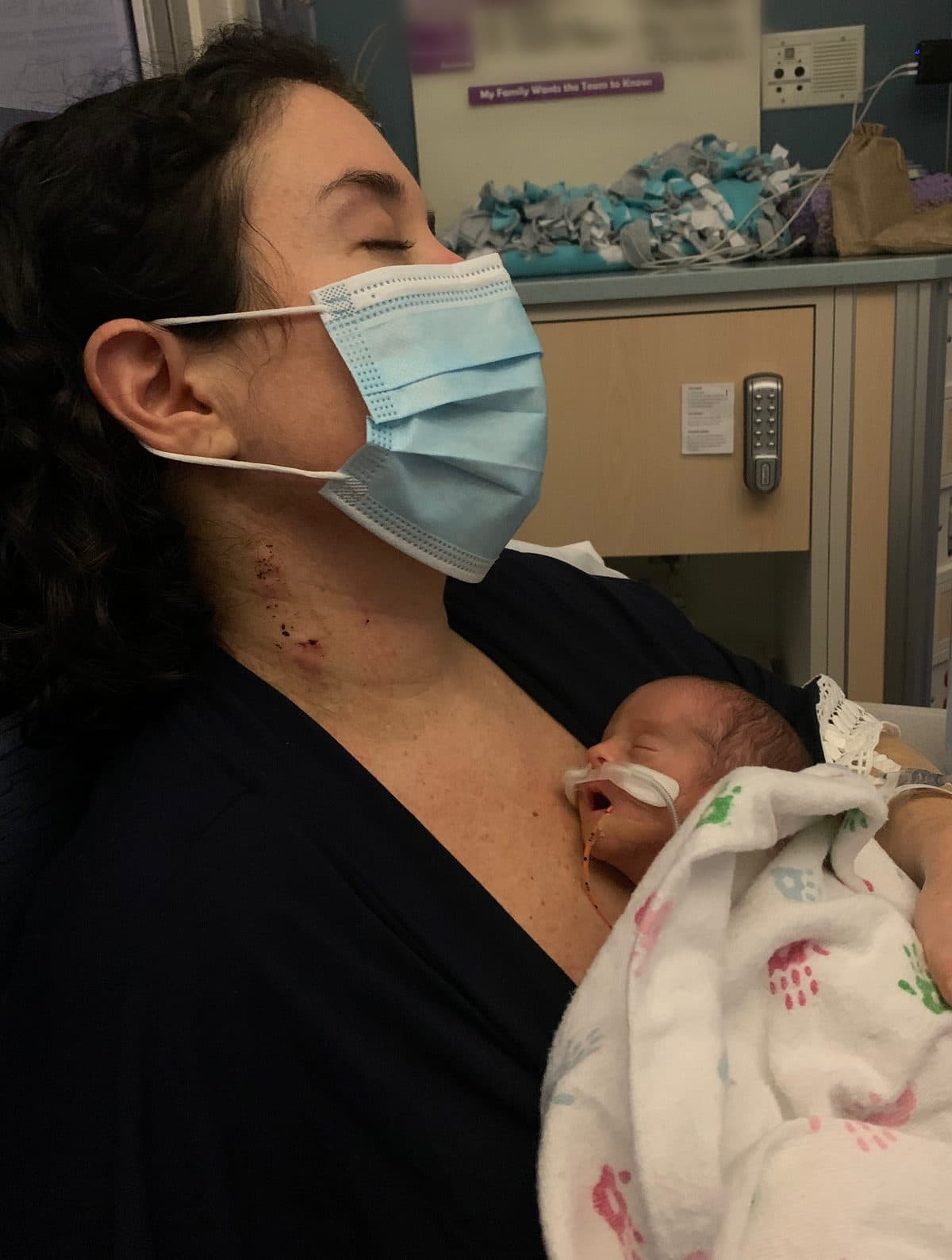 Lauren Rissman, wearing a surgical mask with her eyes closed, holds her newborn daughter against her chest in the NICU. The baby is wrapped in a hospital blanket and has a feeding tube.